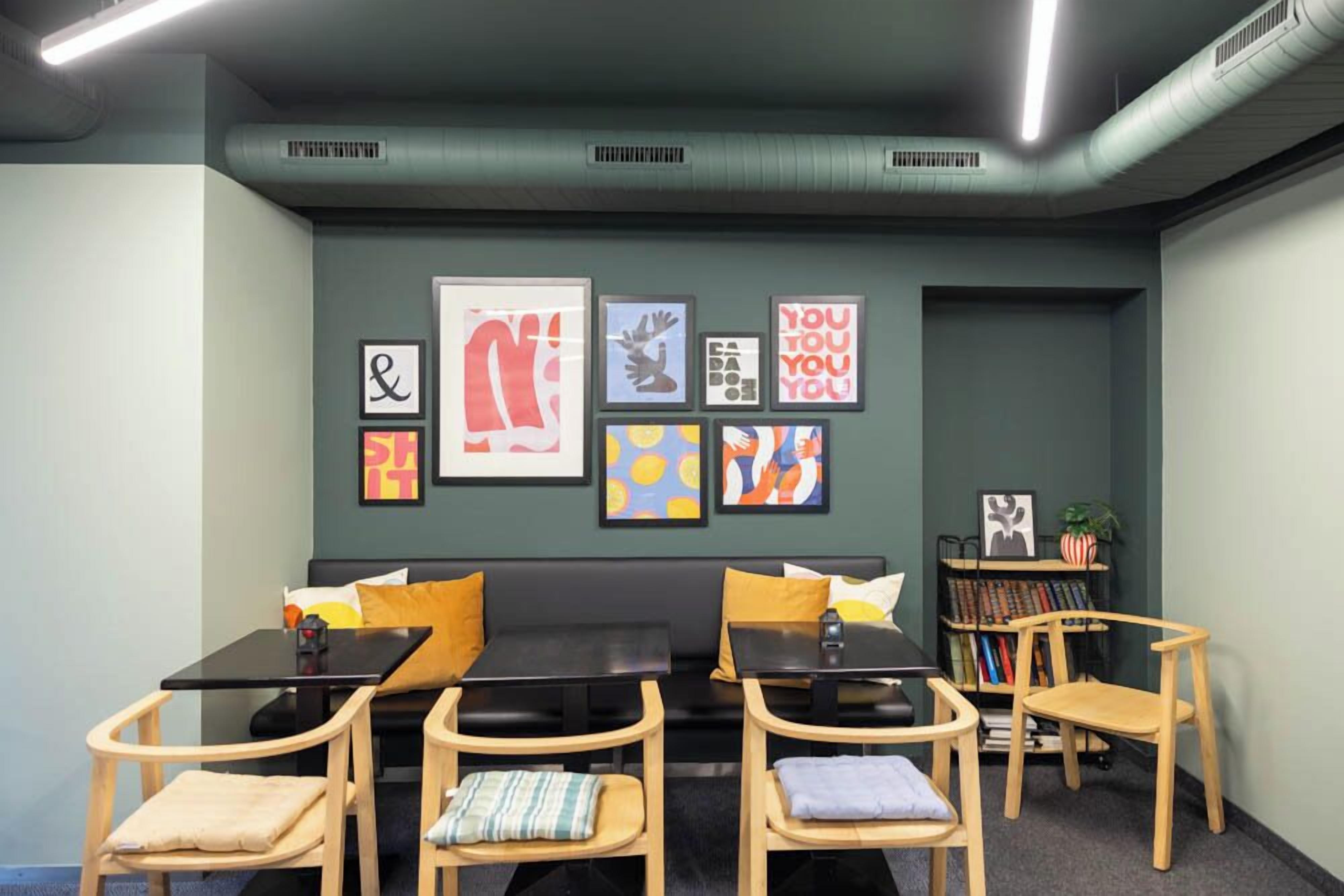 Un área de descanso moderna con un sofá negro, dos mesas y sillas de madera. Hay obras de arte enmarcadas en la pared y una estantería con libros y objetos decorativos.