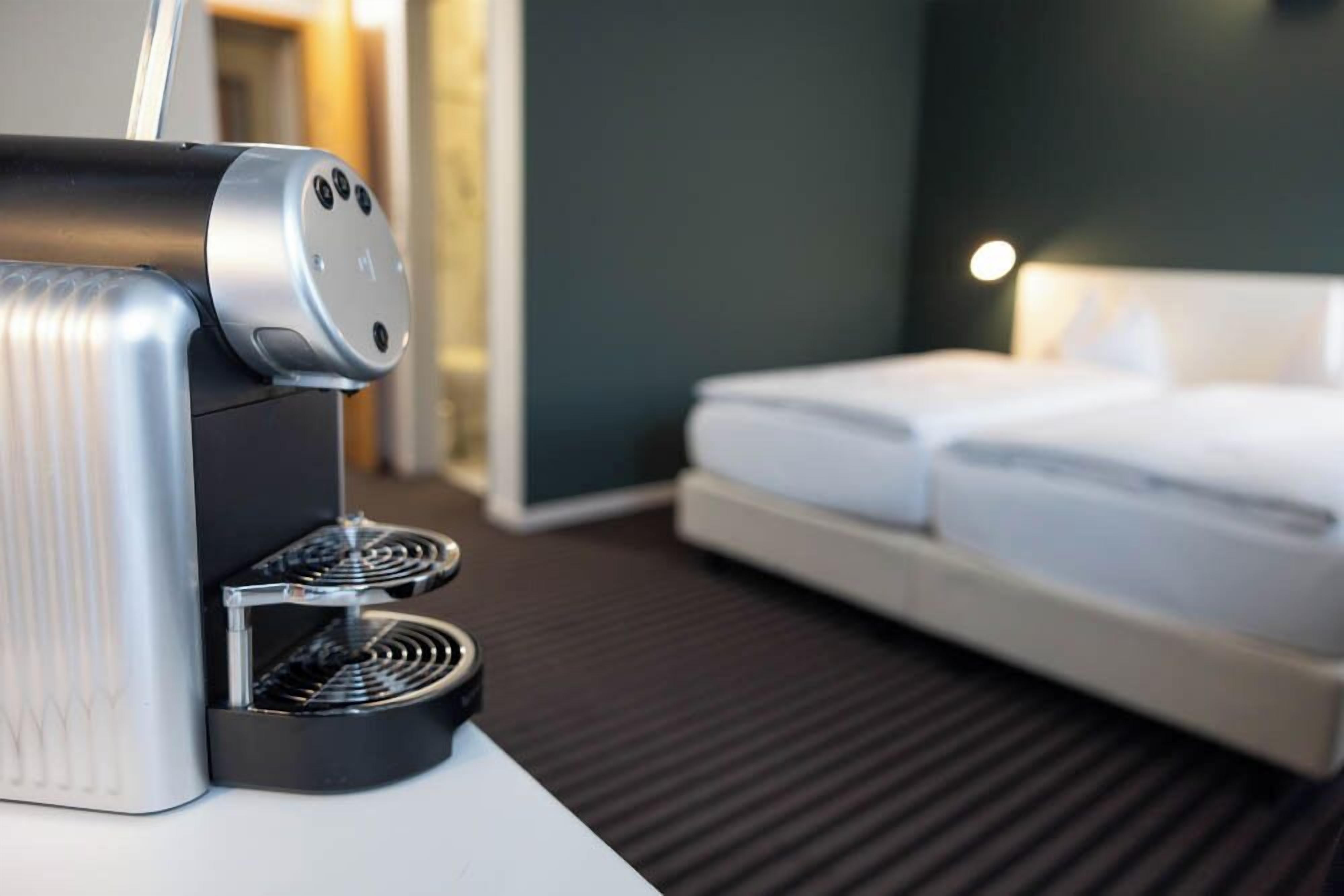 Una habitación de hotel moderna con una cafetera elegante sobre una mesa blanca, una cama bien hecha y una pared de color oscuro.