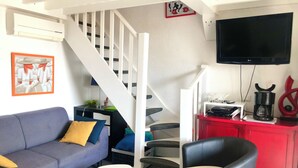 Ein modernes Wohnzimmer mit einem grauen Sofa, einem schwarzen Sessel und einem kleinen weißen Tisch. Eine Treppe mit weißen Geländern führt auf eine höhere Ebene.