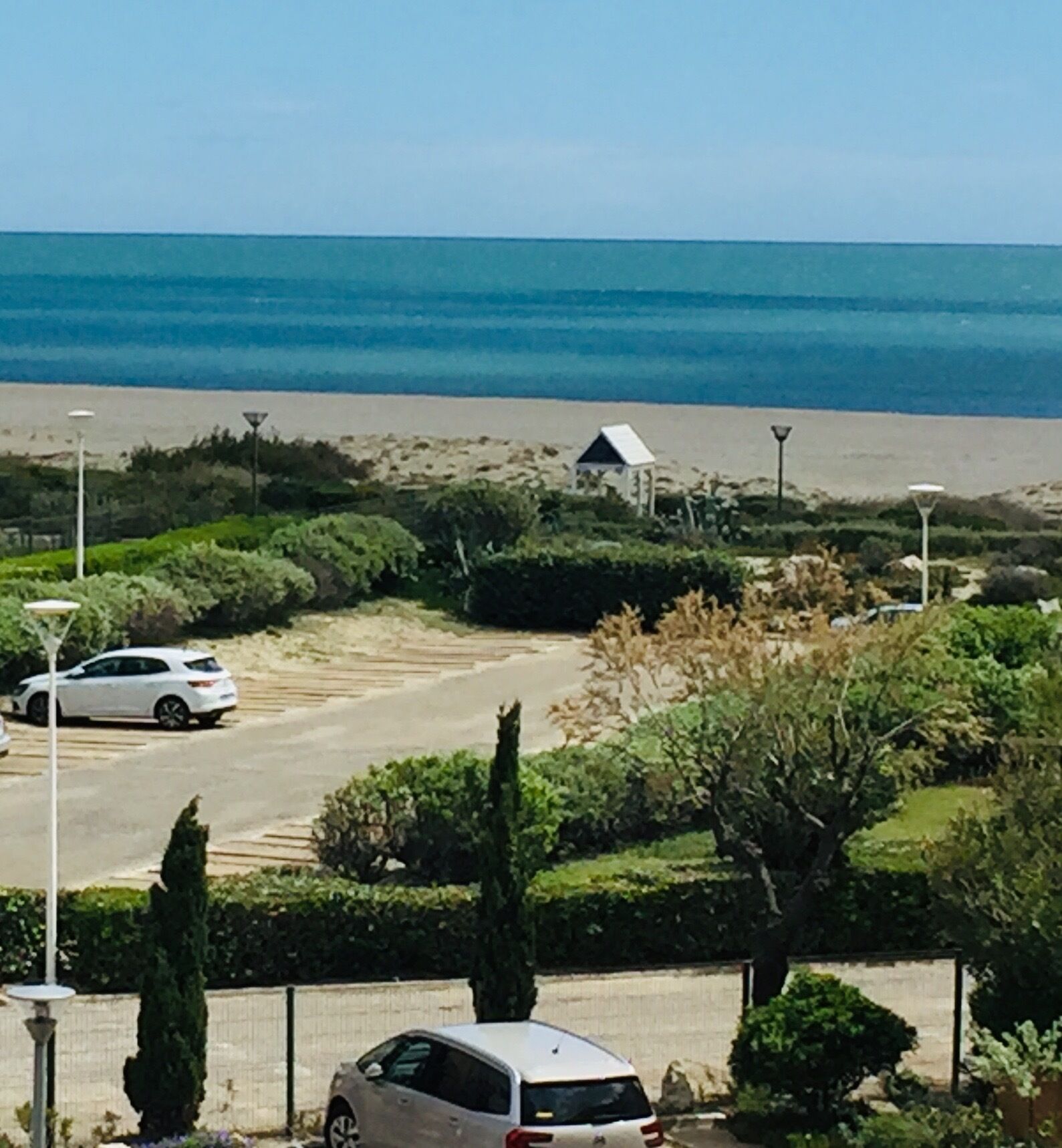 Uma estrada costeira com carros estacionados, um pequeno edifício e uma praia ao fundo.