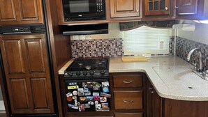 A compact kitchen with wooden cabinets, a microwave, a stove, and a sink. - Spacious Lakeside Travel Trailer: Ultimate Comfort Awaits (Bridgeport)