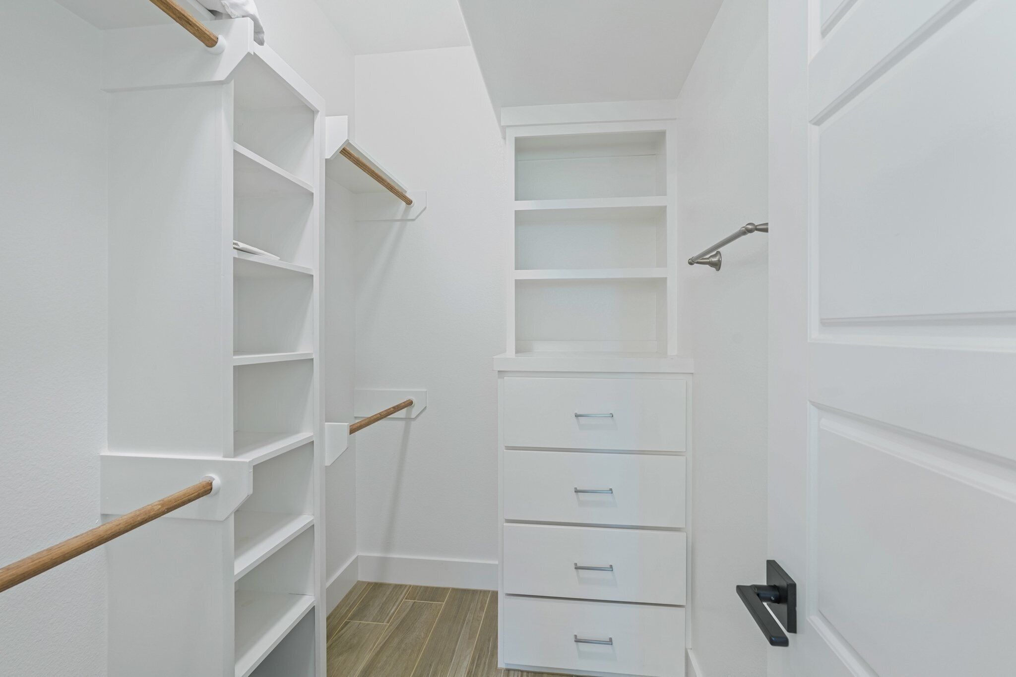 A narrow walk-in closet with white shelving and drawers, a wooden handrail, and a black door handle.