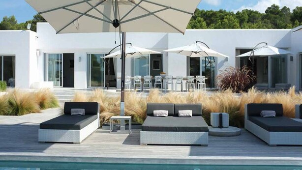A modern outdoor pool area with lounge chairs and umbrellas, surrounded by a wooden deck and a building with large windows. - Gorgeous Villa With Views (Sant Josep de sa Talaia)