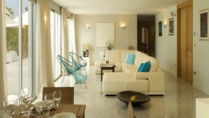 A modern living room with a sofa, a dining table with chairs, and a large window with curtains. - Gorgeous Villa With Views (Sant Josep de sa Talaia)