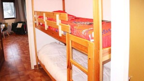 A bunk bed room with a wooden frame, a red mattress, and a white mattress. There is a chair and a window in the room.