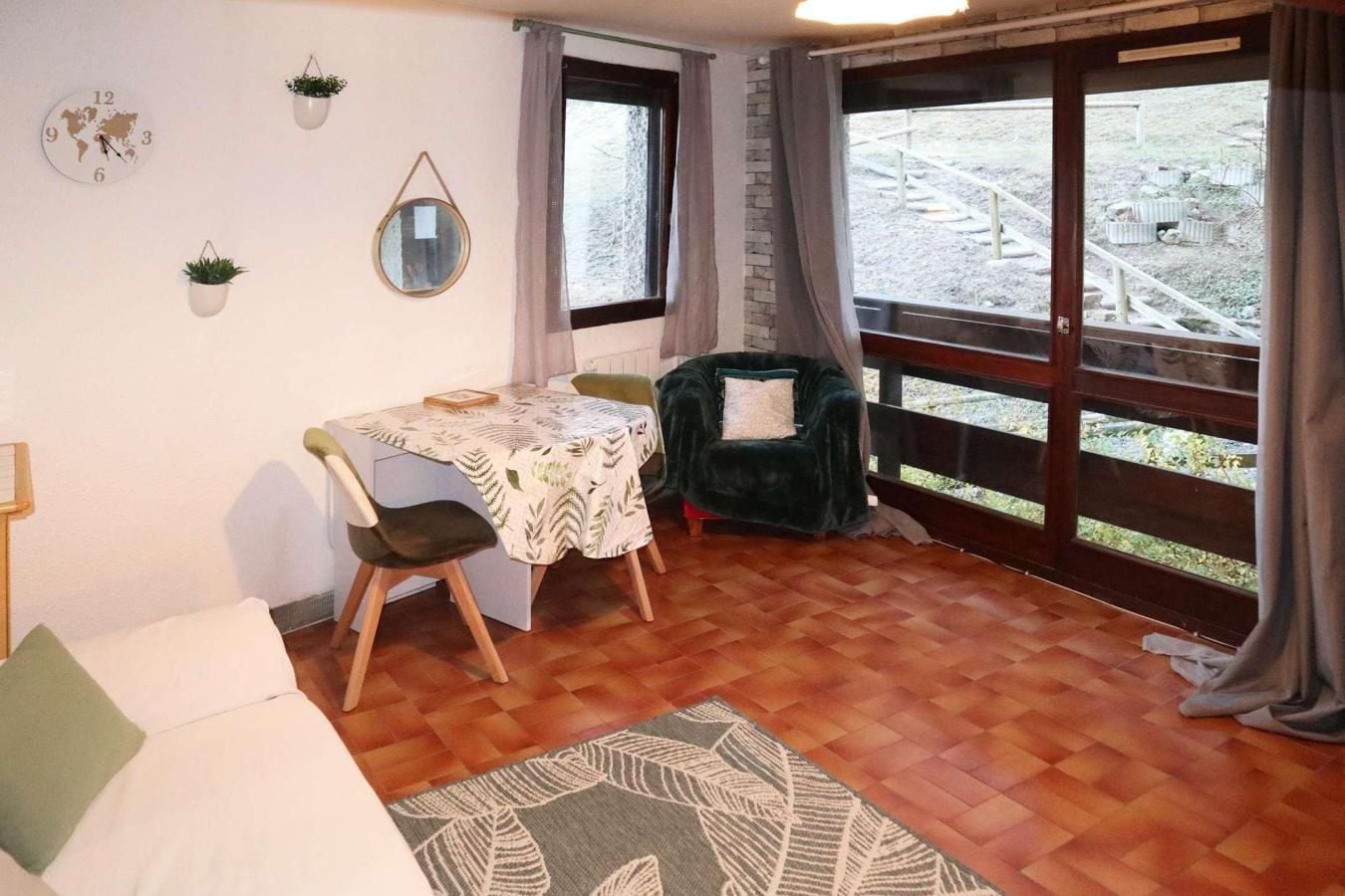 A cozy room with a wooden floor, a dining table with chairs, a green armchair, a world map clock, and a window with a view of a garden.
