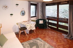 A cozy room with a wooden floor, a dining table with chairs, a green armchair, a world map clock, and a window with a view of a garden. - 50m from ski slopes, 1st floor, 20m², Les Orres (Les Orres)