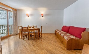 A room with a wooden dining table and chairs, a wooden sofa with red cushions, and two wall-mounted lamps.