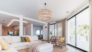 A modern living room with a white sofa, a beige ottoman, and a large window with a view of palm trees. - Superb Villa With Private Lagoon Pool (Javea)