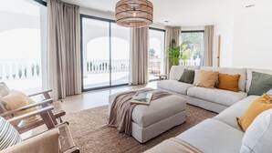 A spacious living room with a large sofa, a coffee table, and a wooden chair. There is a large window with curtains, a hanging light fixture, and a plant. - Superb Villa With Private Lagoon Pool (Javea)