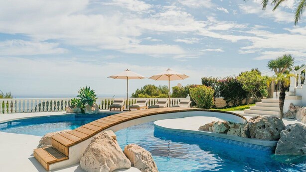 A pool with a curved design, surrounded by lounge chairs and umbrellas, with a view of the ocean. - Superb Villa With Private Lagoon Pool (Javea)