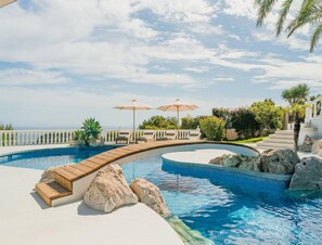 A pool with a curved design, surrounded by lounge chairs and umbrellas, with a view of the ocean.