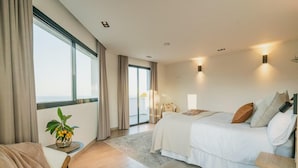 A modern bedroom with a large bed, a chair, a small table with a plant, and a view of the outdoors through a sliding door. - Superb Villa With Private Lagoon Pool (Javea)