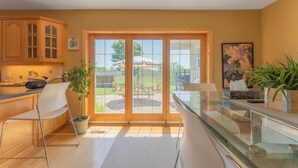 A modern kitchen with wooden cabinets, a glass dining table, and a view of the backyard through large sliding doors.