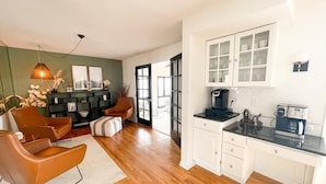 A modern kitchen with white cabinets, a black countertop, and a coffee machine. There is a seating area with a brown chair and a rug.