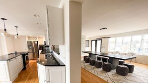 A modern kitchen with white cabinetry, stainless steel appliances, and a black countertop. There is a dining area with a large table and chairs, and a view of the city through large windows.