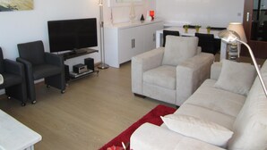 A living room with a flat-screen TV, a sofa, a coffee table, and a red rug. There are two paintings on the wall, a lamp, and a cabinet. - Apartment in Blankenberge With Sea View (Blankenberge)