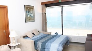 A hotel room with a bed, a sofa, a table with a lamp, a painting on the wall, and a large window with a view of the beach. - Apartment in Blankenberge With Sea View (Blankenberge)