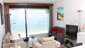 A modern living room with a large window overlooking the sea, a flat-screen TV, and a dining area. - Apartment in Blankenberge With Sea View (Blankenberge)