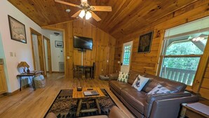 Interior acogedor de una cabaña con techo de madera, sofá de cuero, televisor y un amplio ventanal con vistas a árboles.