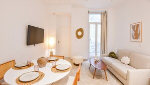 A modern living room with a round dining table set for four, a sofa, a TV mounted on the wall, and a window with curtains.