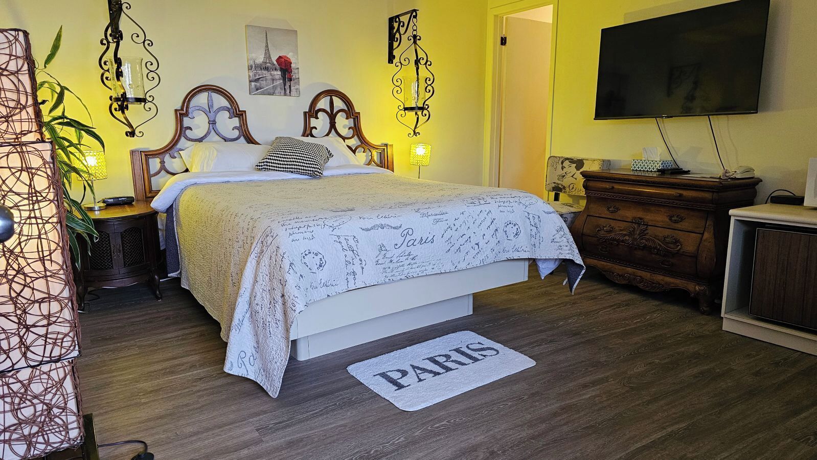 A bedroom with a bed, bedside tables, a dresser, a television, and a rug with 'PARIS' on it.