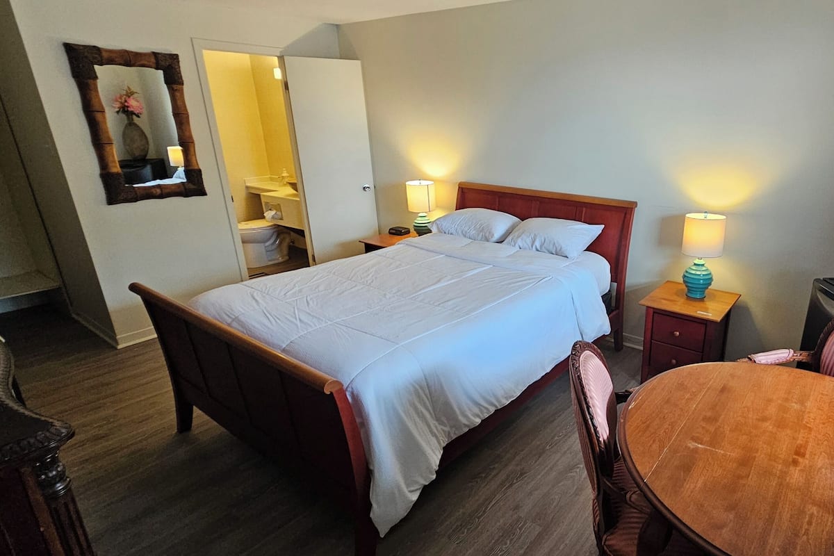Une chambre d’hôtel avec un lit, des tables de chevet, une chaise, une petite table, un miroir et une fenêtre.