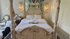 Una cama con dosel con una colcha blanca y una almohada con forma de cisne. Hay apliques de pared con flecos, un espejo y una mesita con una lámpara y adornos.