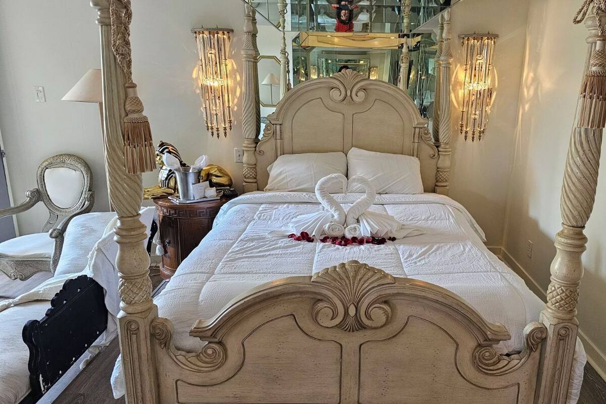 Un lit à baldaquin avec un couvre-lit blanc et un oreiller en forme de cygne. On y trouve des appliques murales ornées de glands, un miroir et une petite table avec une lampe et des objets décoratifs.