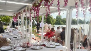Un espacio al aire libre con mesas dispuestas para un evento formal, rodeado de vegetación y una masa de agua.