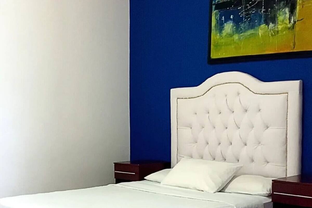 Uma cama bem arrumada com cabeceira branca e travesseiros, mesa de cabeceira de madeira e uma pequena mesa.