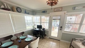 A dining area with a wooden table, four chairs, and blue plates. There is a desk with a computer and a lamp. The room has a door leading outside, and windows with blinds.