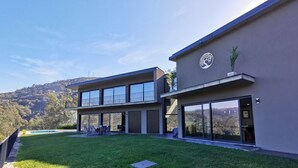 Une maison moderne à deux étages, dotée de grandes fenêtres en verre, d’une pelouse bien entretenue et offrant une vue sur un paysage vallonné.