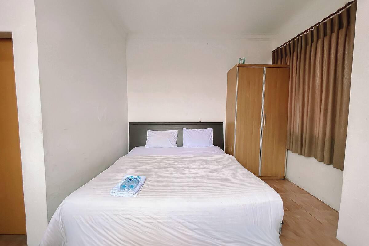 Una cama individual con ropa de cama y almohadas blancas, un armario de madera y una ventana con cortinas marrones.