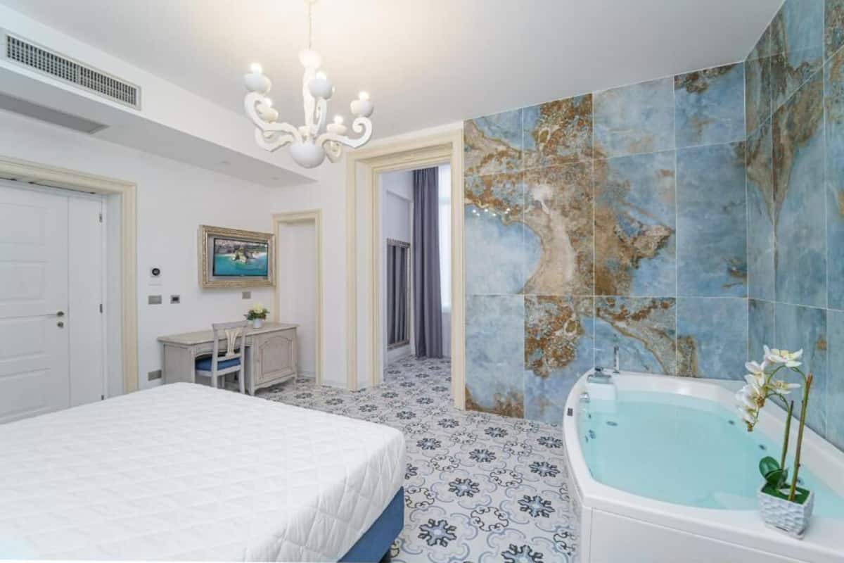 A bedroom with a large bed, a vanity with a mirror, and a bathtub with a flower vase.
