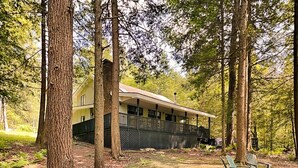 A cabin with a porch, surrounded by tall trees and a grassy area with Adirondack chairs. - Koi at the Lake by Taylor Made Deep Creek (Oakland)