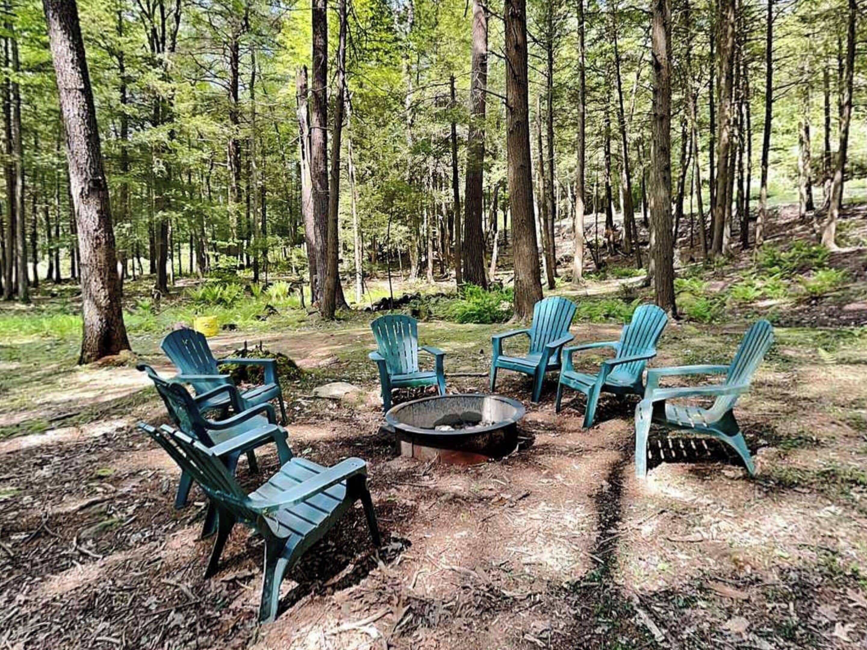 A forest setting with a fire pit and several Adirondack chairs arranged around it.