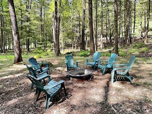 A forest setting with a fire pit and several Adirondack chairs arranged around it. - Koi at the Lake by Taylor Made Deep Creek (Oakland)