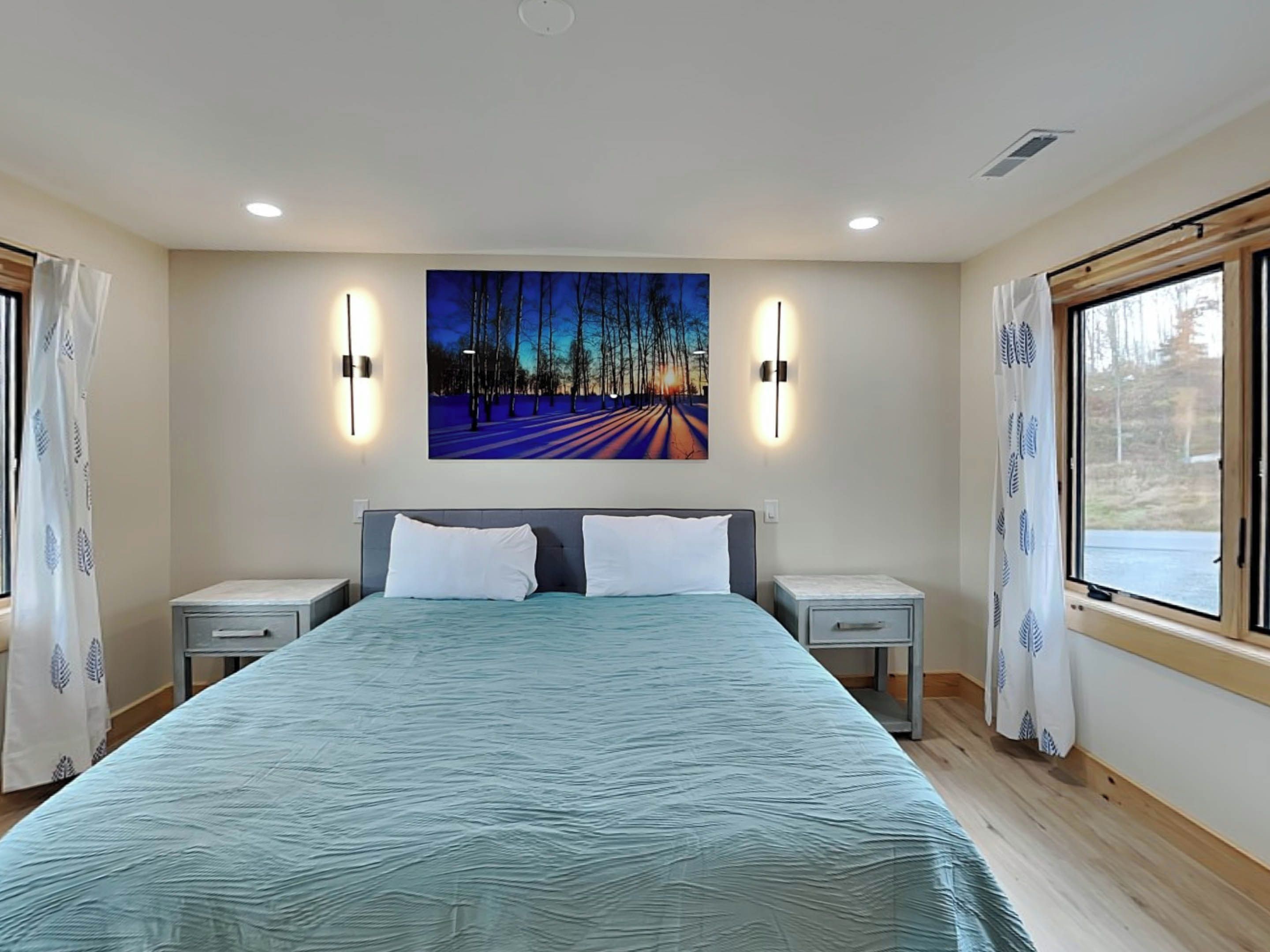 A bedroom with a large bed, two bedside tables, a nightstand, a painting on the wall, and a window with curtains.