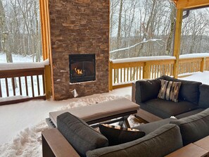 Un rincón acogedor al aire libre con una chimenea de piedra, una terraza de madera y vistas a un bosque nevado.