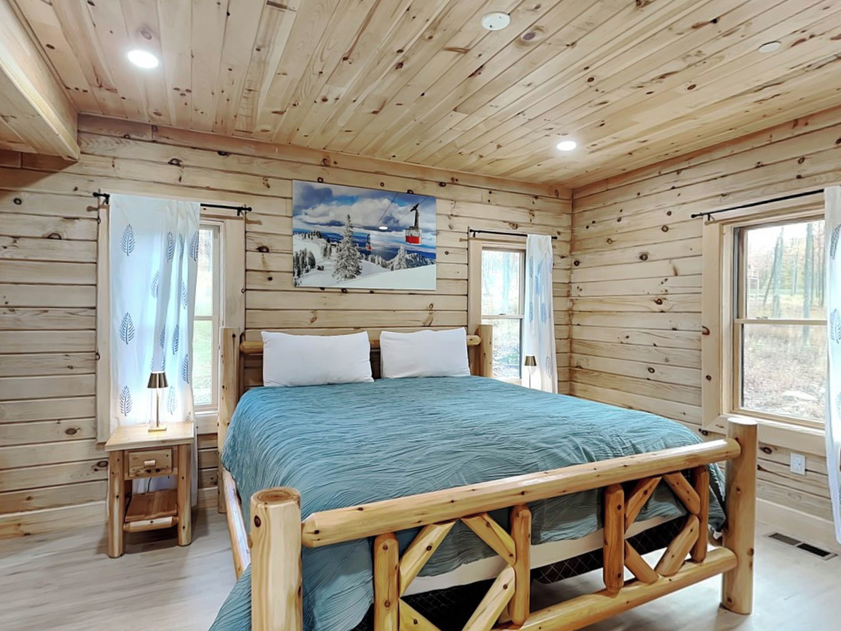 A wooden cabin bedroom with a bed, a nightstand, and a painting of a snowy landscape.