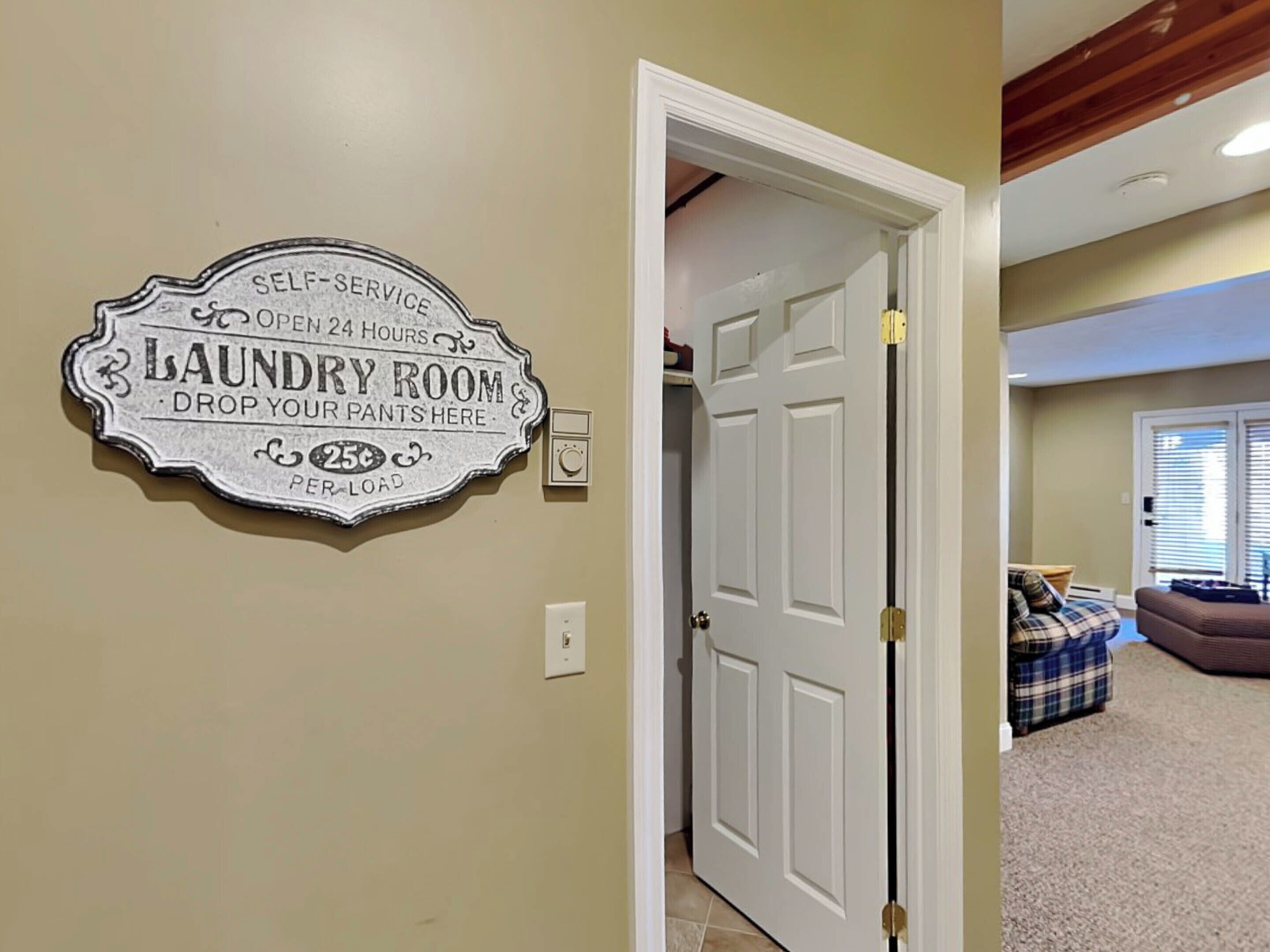 A laundry room sign with self-service and 24-hour availability, a door leading to a room with a blue plaid couch and a window with blinds.