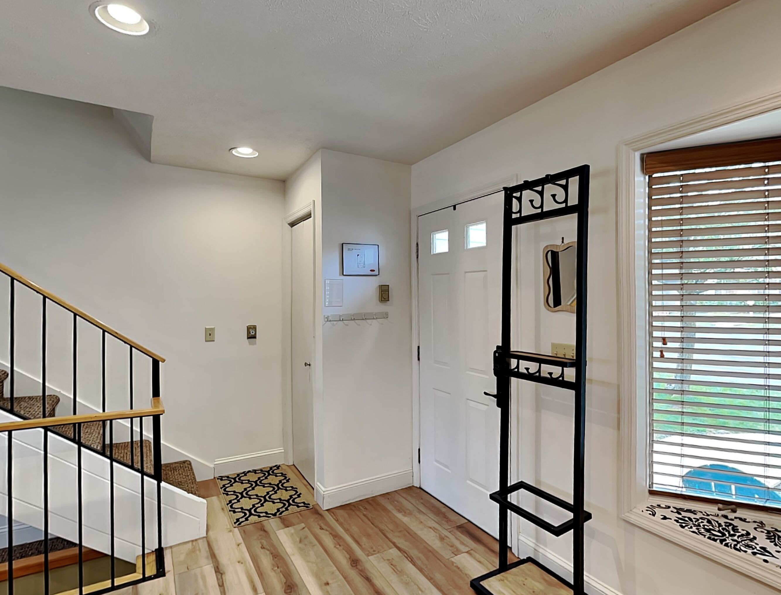 A hallway with a wooden staircase, a door with a mirror, and a window with blinds.
