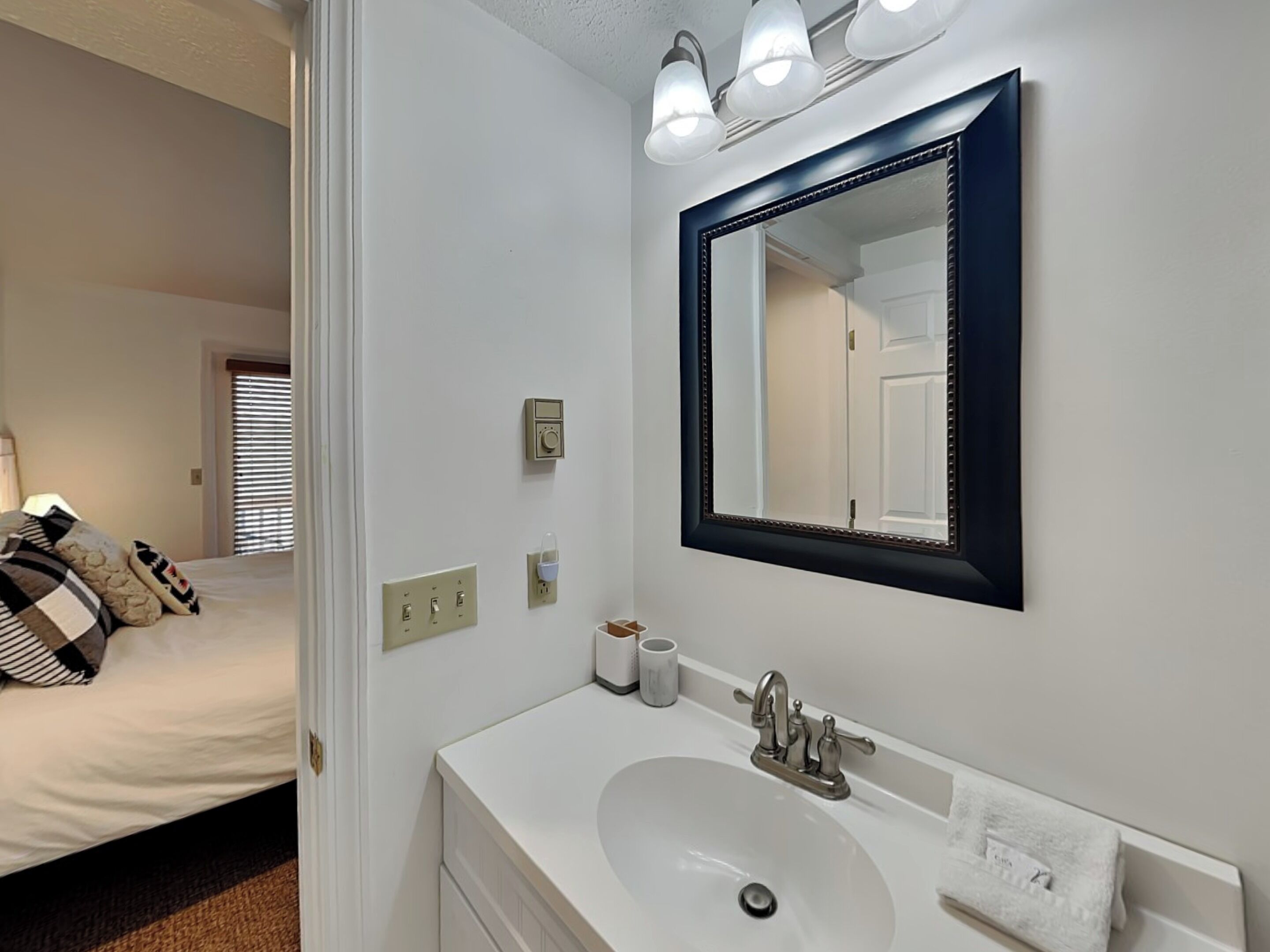 A hotel bathroom with a white sink, a large mirror, and a bed visible through an open door.