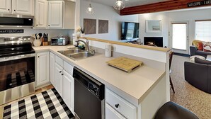 A modern kitchen with white cabinets, a stainless steel range, and a dishwasher. There is a wooden island with a cutting board, a sink, and a countertop with various kitchen items. A television is mounted above the counter, and there is a fireplace with a mantel. - Happy Time by Taylor Made Deep Creek (McHenry)