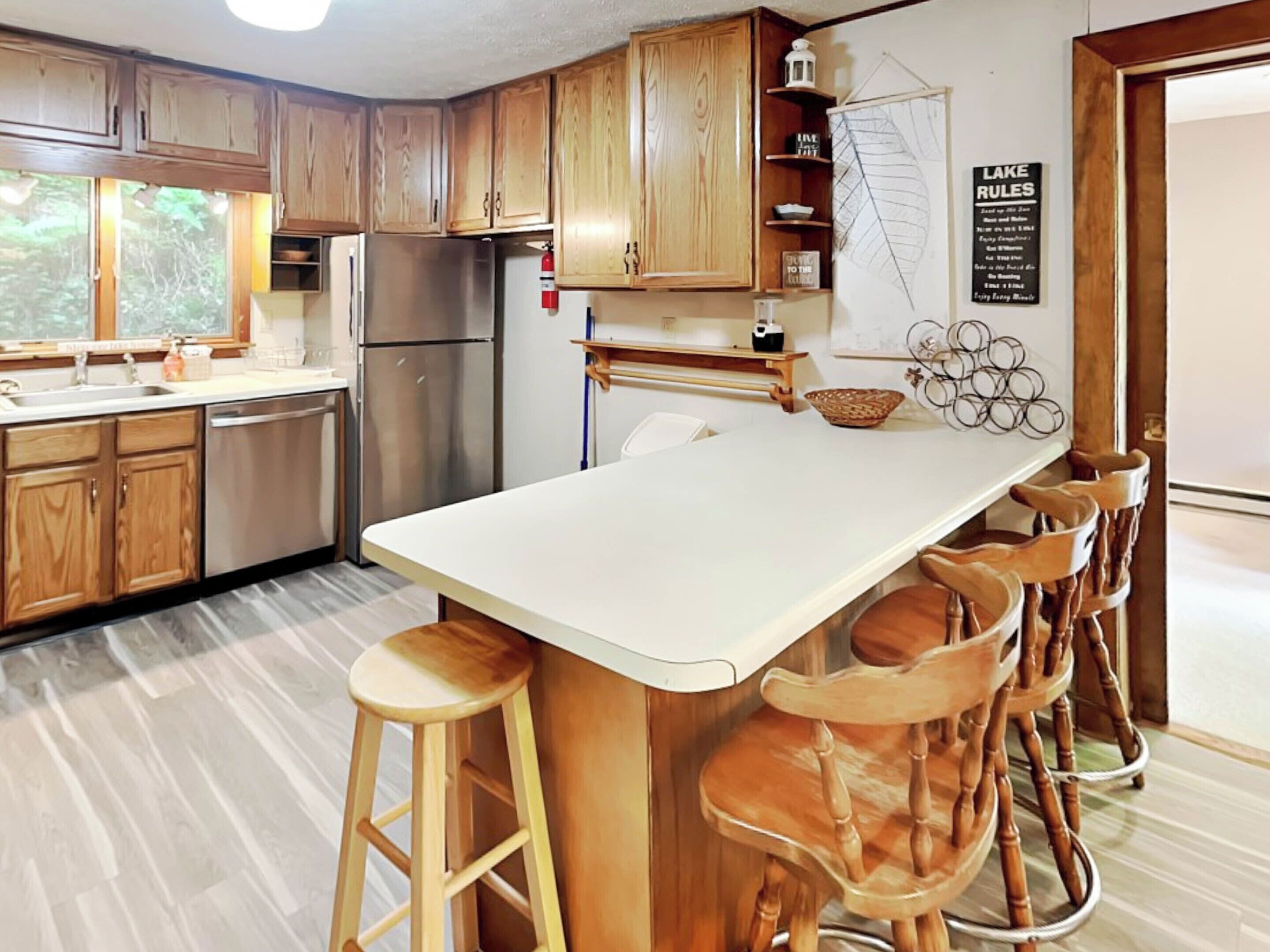 A kitchen with wooden cabinets, a stainless steel refrigerator, a white countertop, and a bar area with stools.