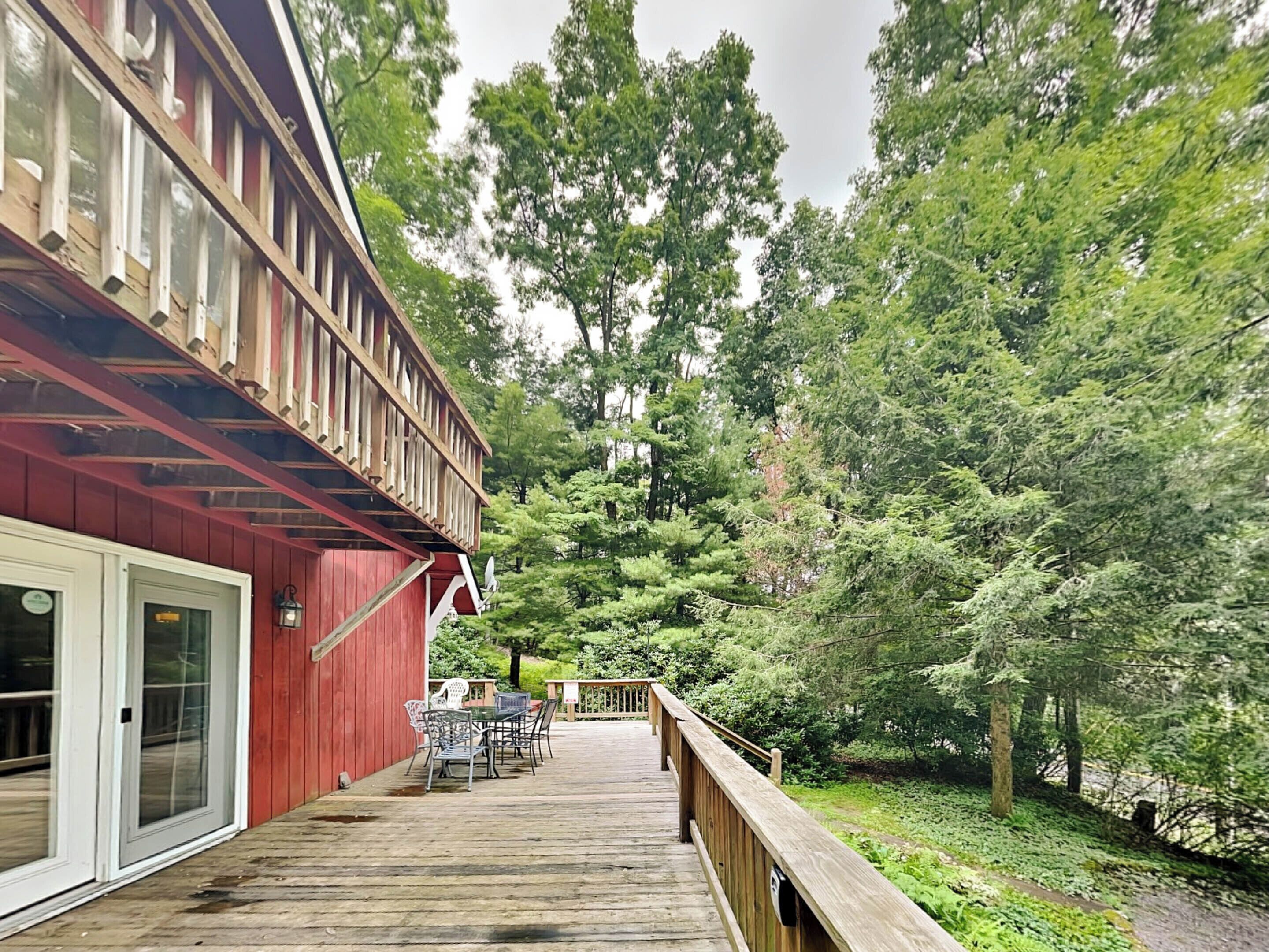Une terrasse en bois avec des meubles d’extérieur, un bâtiment rouge doté d’un balcon, et une zone boisée en arrière-plan.