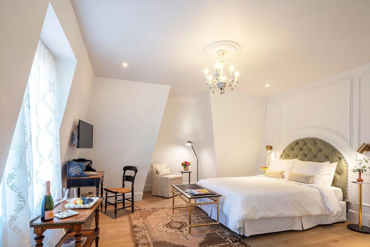 A bedroom with a large bed, a desk with a chair, a small table, a chandelier, and a television on the wall.