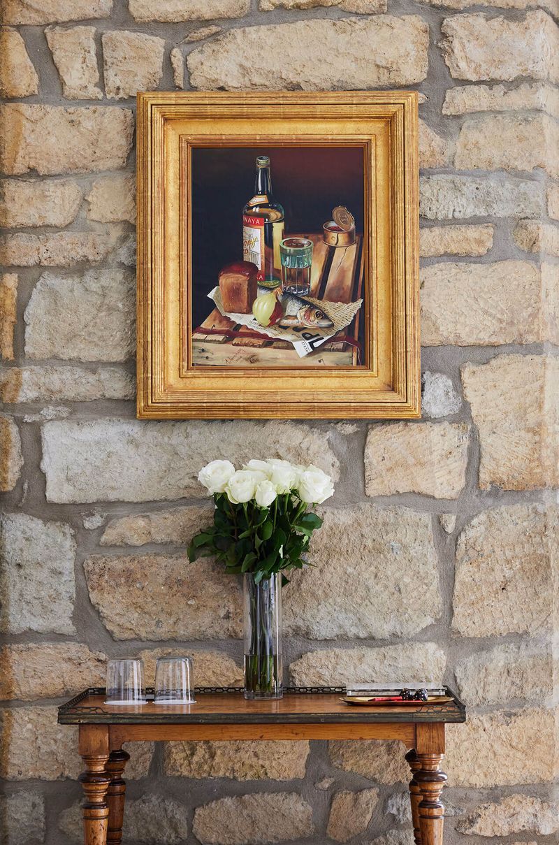 A framed painting of a still life on a stone wall, a vase of white flowers, and a wooden table with glasses and a book.
