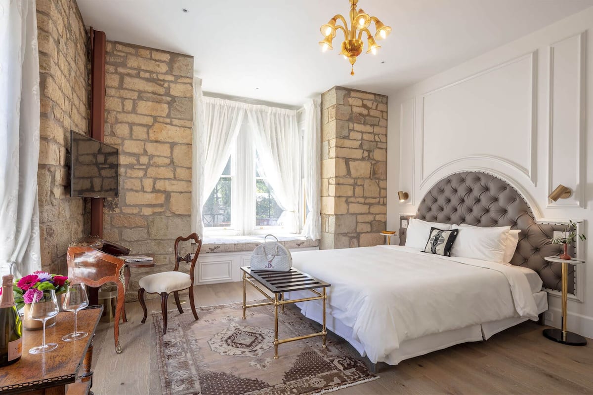 A bedroom with a stone wall, a large bed, a chandelier, and a window with curtains.